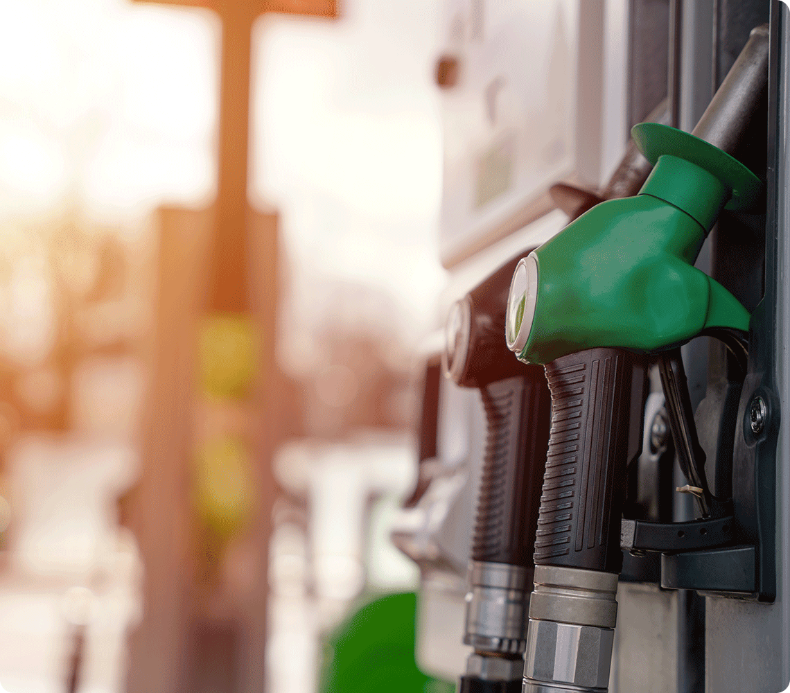 Close-up of a green fuel pump nozzle at a gas station with a blurry background of another pump and sunlight.